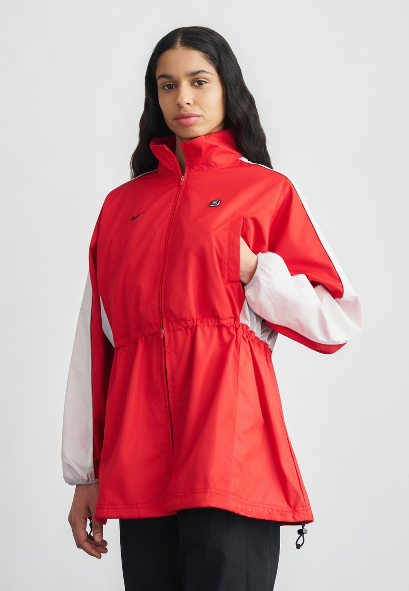 Jachetă roșie de tip windbreaker cu mâneci albe, talie cu șnur și guler înalt. Are un logo Nike pe piept și un fermoar parțial deschis în față.