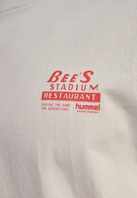 Logotip in besedilo na svetli tkanini: "Bee's Stadium Restaurant", "Postrežemo igro skozi generacije" in "hummel sportswear" v rdeči barvi.