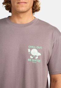 Szürke pamut póló, amelynek elülső grafikáján a "CHILL OUT. BE WATER" felirat aqua színben látható, egy rajzfilmfigurát ábrázol, aki egy surf deszkát tart.