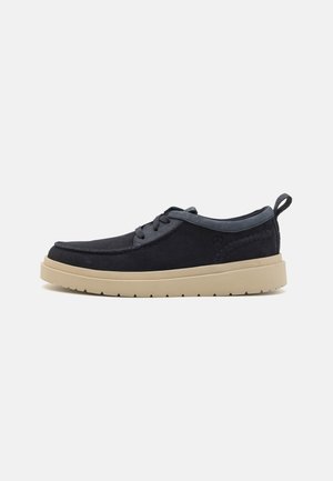 Pantofii din suede bleumarin închis au o talpă plată din cauciuc bej, vârful rotund și un design cu șireturi. Dispun de un detaliu decorativ cu cusătura pe lateral.