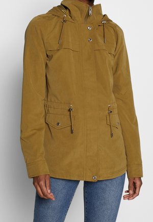 Chaqueta marrón con capucha, cremallera frontal, botones a presión, cintura con cordón, bolsillos con solapa, llevada con vaqueros azules en una persona con fondo liso.