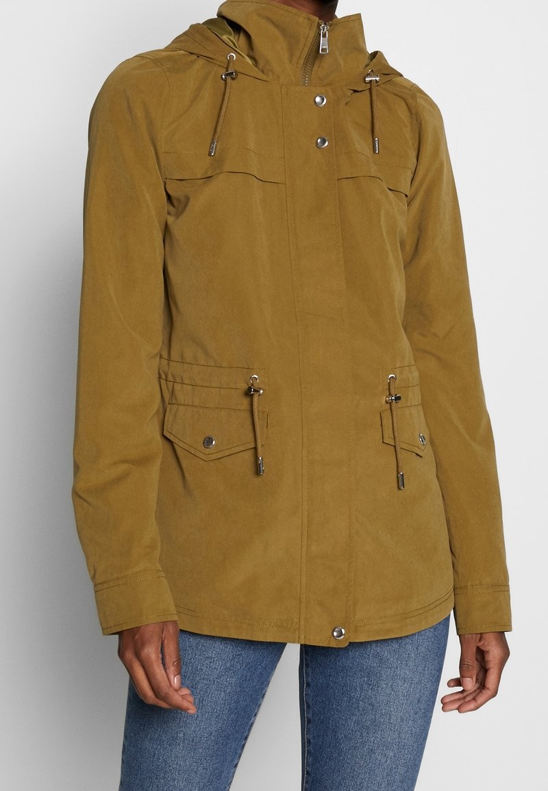 Veste à capuche marron avec fermeture éclair devant, boutons-pression, taille à cordon de serrage, poches à rabat, portée avec un jean bleu sur une personne devant un fond uni.