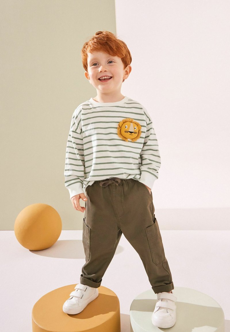 Enfant roux souriant debout sur des plateformes rondes portant une chemise rayée avec un écusson lion, un pantalon marron et des baskets blanches dans un studio lumineux.