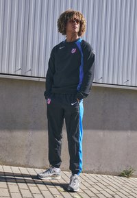 Sort sweatshirt med blå detaljer, prydet med et logo, sammen med sorte bukser med en blå stribe. Modellen har grå sneakers og solbriller på.