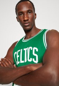 Grünes Basketball-Trikot mit weißem "CELTICS"-Text, Netzstoff, runder Halsausschnitt und kontrastierenden Akzenten, getragen von einem Modell mit verschränkten Armen.