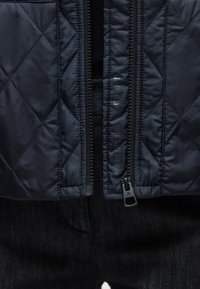 Lähikuva mustasta tikatusta takista, jossa osittain vetoketjulla suljettu vetoketju, jonka saumassa lukee "MARCO POLO X FUNCTION".