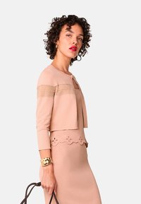 Beige gestrickte Strickjacke mit langen Ärmeln, mit einem Schlüsselloch-Ausschnitt und gestreiftem Design am Oberteil. Kombiniert mit einem passenden Midikleid.