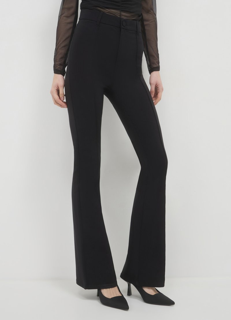 Schwarze Schlaghosen mit glatter Textur, ausgestattet mit einer Taillenbetonung und Seitennähten, ergänzt durch spitze schwarze Absatzschuhe.