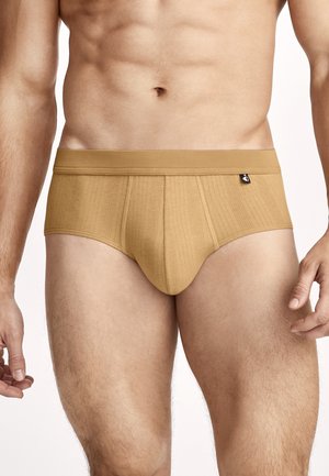 Fit male torso wearing tan ribbed brief-style underwear with a small black tag on the waistband against a neutral background.