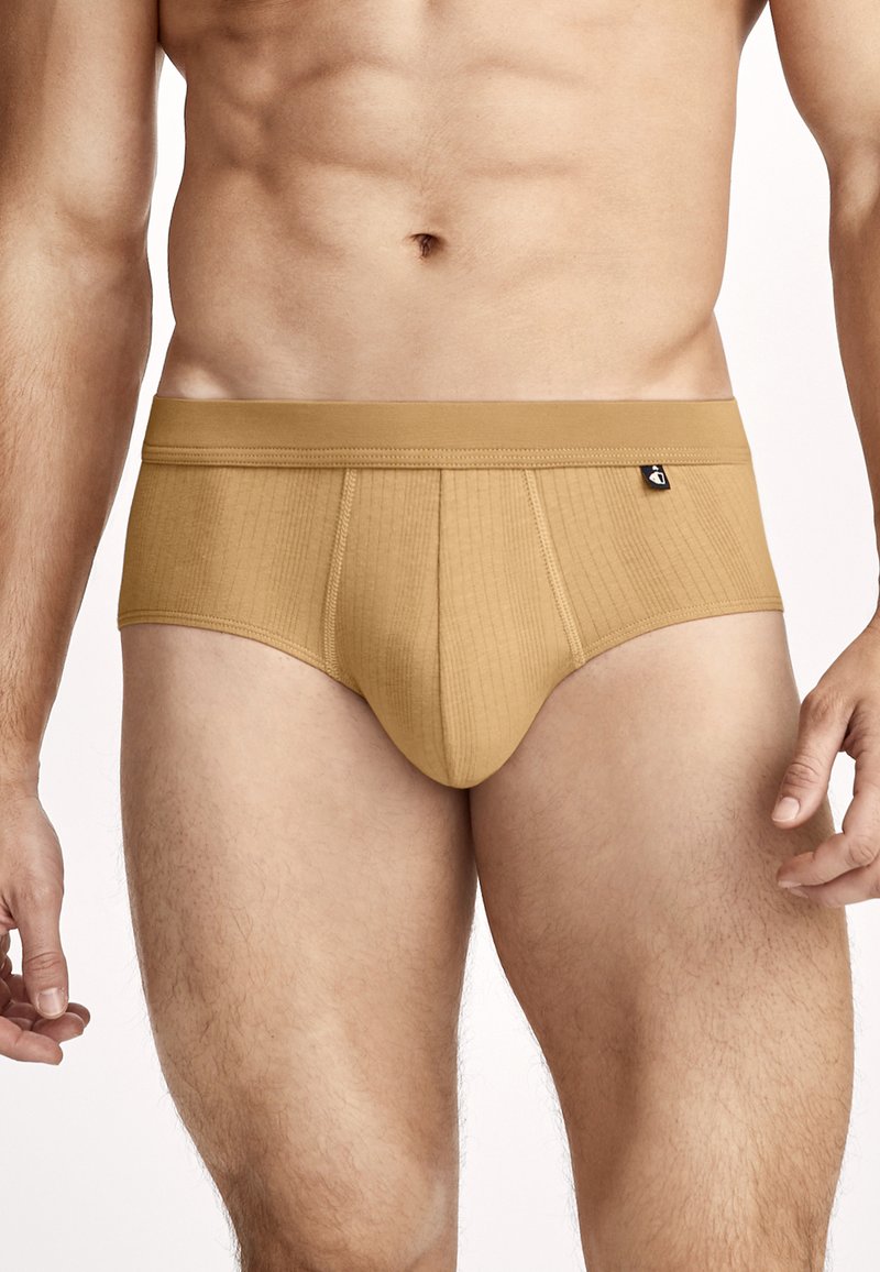 Athletischer männlicher Oberkörper, der beige gerippte Unterhose im Slip-Stil mit einem kleinen schwarzen Etikett am Bund vor neutralem Hintergrund trägt.