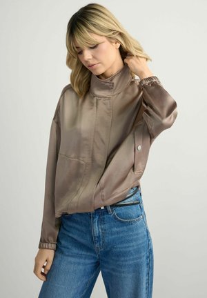 Blondynka poprawiająca kołnierz luźnej brązowoszarej satynowej kurtki, nosząca niebieskie dżinsy z wysokim stanem, stojąca na tle jednolitego jasnego tła.