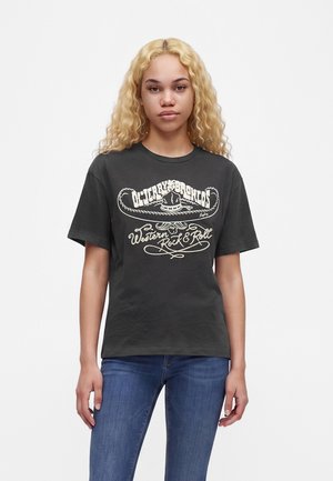 Giovane donna con lunghi capelli biondi ricci che indossa una t-shirt nera con grafica e jeans blu su uno sfondo bianco uniforme.