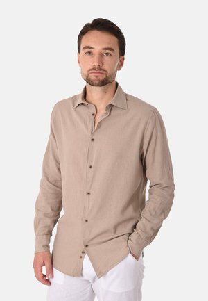 Uomo con capelli corti scuri e barba, che indossa una camicia beige a maniche lunghe con bottoni e pantaloni bianchi su uno sfondo chiaro e uniforme.