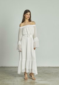 Weißes schulterfreies langes Kleid mit Rüschensäumen und tailliertem Gürtel. Texturiertes Gewebe, gestufter Saum, kombiniert mit lässigen metallischen Absatzsandalen.
