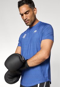 Μπλε αθλητική μπλούζα με γεωμετρικό μοτίβο, κοντά μανίκια και μαύρα γάντια πυγμαχίας. Λείο ύφασμα, εφαρμοστή σχεδίαση.