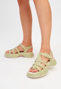 Gröna syntetiska sandaler med korsade remmar, öppen häl, justerbar ankle buckle och en tjock sula med en böjd design.