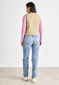 Tan ermeløs vest, ribbet rosa langermet skjorte, lyseblå dong jeans, og hvite klumpete joggesko. Uformelt antrekk sett fra bak.