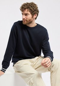 Pull en col rond à manches longues bleu marine avec des poignets côtelés, mettant en avant trois boutons décoratifs sur l'épaule gauche. Porté avec un pantalon beige clair.