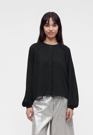 Jeune femme aux longs cheveux foncés portant un chemisier noir ample à manches longues et un pantalon large argenté brillant, debout devant un fond uni.