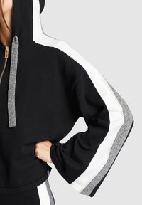 Personne portant un sweat à capuche noir avec des rayures blanches et grises sur les manches et des cordons gris, main dans la poche, sur fond clair uni.