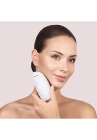 Appareil de nettoyage facial blanc avec un design courbé, une texture en silicone et des boutons, tenu contre le visage d'une personne ayant la peau lisse.