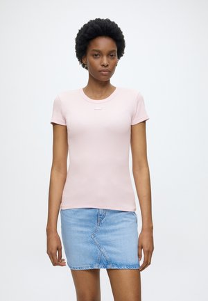 Nő világos rózsaszín rövid ujjú pólót és világoskék farmer miniszoknyát visel, sima fehér háttér előtt áll.