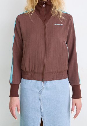 Kobieta w brązowej kurtce na zamek Adidas z jasnoniebieskimi paskami na rękawach, w połączeniu z jasnoniebieską spódnicą dżinsową z rozcięciem z przodu.