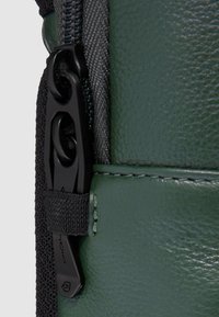 Detail van een groene leren tas met een zwarte rits en een stoffen riem. De textuur is glad met fijne stiknaden, waarbij functionele accenten worden benadrukt.