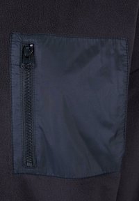 Tessuto in pile nero con una tasca in nylon liscio dotata di zip sul lato sinistro. Contrasti di texture tra il pile e il nylon lucido.