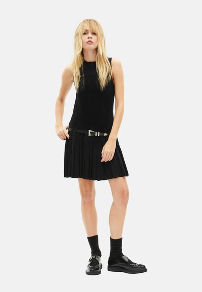 Vestito nero plissettato con corpetto aderente, scollo rotondo e cintura nera con fibbia argentata, abbinato a scarpe e calzini neri.