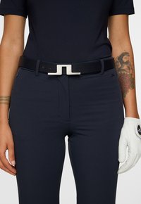 Persona che indossa pantaloni e camicia blu scuro, cintura nera con fibbia argentata, braccio tatuato e guanto bianco sulla mano destra.