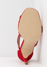 Talons en suede rouge avec un bout pointu, des lanières à la cheville et un fin stiletto. La semelle est beige avec des rainures horizontales.