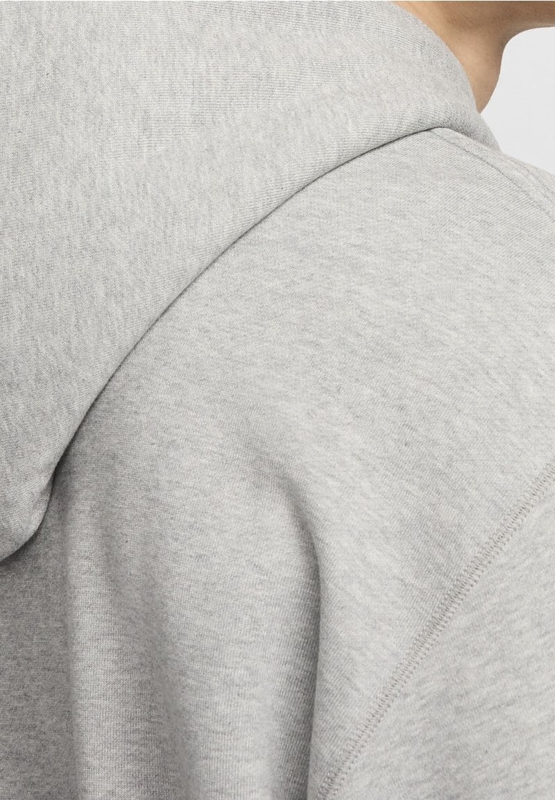 Detailní záběr na rameno osoby, která má na sobě světle šedou mikinu s kapucí, viditelným šitím a měkkou texturou látky.