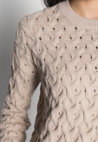 Béžový pletený svetr s texturovaným kabelovým vzorem, kulatým výstřihem a viditelnými stehy po celé tkanině.