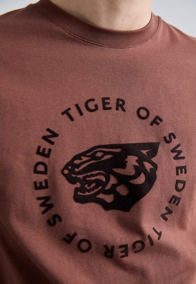 Brūns kokvilnas T-krekls ar melnu tīģera grafiku un tekstu "TIGER OF SWEDEN" apļa izkārtojumā apkārt grafikam. Īsas piedurknes.