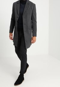 Cappotto grigio di lana con chiusura a un bottone e rever a lancia, indossato sopra un dolcevita blu navy a trama e pantaloni neri, abbinato a scarpe nere.