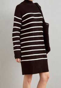 Robe-pull tricotée marron foncé avec des rayures horizontales blanches, munie d'un col montant et de manches longues, associée à un sac texturé noir.