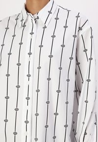 Chemise blanche boutonnée avec un motif répétitif de logos noirs. Conçue avec un col et des manches longues, fabriquée dans un tissu lisse.