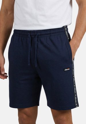 Uomo che indossa pantaloncini blu navy con coulisse con nastro logo sui lati e piccolo patch logo sulla coscia sinistra, abbinato a una maglietta bianca.