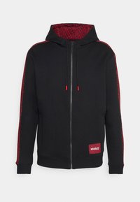 Sudadera negra con cremallera, interior rojo estampado y bandas de acento. Presenta cordones rojos y un parche de logo en el dobladillo.