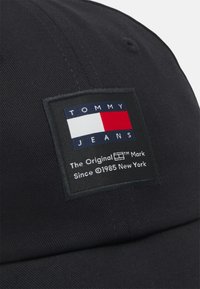 Gorra negra con un parche rectangular de logo que presenta colores rojo, blanco y azul. Incluye "TOMMY JEANS" y detalles de texto en blanco.