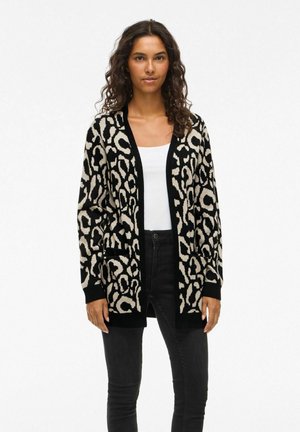 Gilet tricoté avec un imprimé léopard noir et beige, à design ouvert, manches longues et poches, associé à un jean noir et un haut blanc.