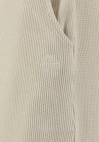 Gros plan sur un tissu beige en maille gaufrée avec un subtil logo brodé en forme de cercle et de croix près d'une couture en V.