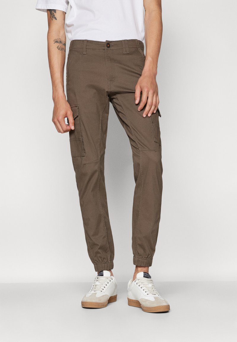 Pantalones cargo marrones con puños elásticos, bolsillos laterales y cierre de botón. Combinados con zapatillas blancas y una camiseta blanca lisa.