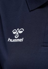 Námořnická modrá polo košile s texturovanou tkaninou. Na levé části hrudi se nachází bílé logo hummel, což zdůrazňuje její sportovní design.