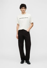 Νεαρός άνδρας που στέκεται φορώντας λευκό t-shirt με λογότυπο Emporio Armani, μαύρο παντελόνι και μαύρα παπούτσια, με το ένα χέρι στην τσέπη.