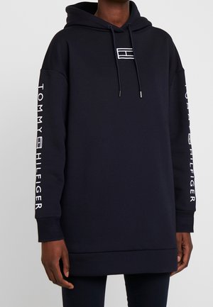 Person iført en sort Tommy Hilfiger hættetrøje med hvidt logo på brystet og tekst på begge ærmer, stående mod en ensartet lys baggrund.