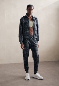 Mørkegrå treningsdrakt med leopardmønster, som har en glidelåsjakke og bukser med elastisk midje, kombinert med hvite joggesko.