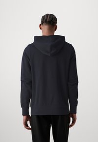 Tmavě modrá mikina s kapucí vyrobená z měkké tkaniny, má klokaní kapsu a kapuci s šňůrkou. Elastické manžety a lem dokončují design.