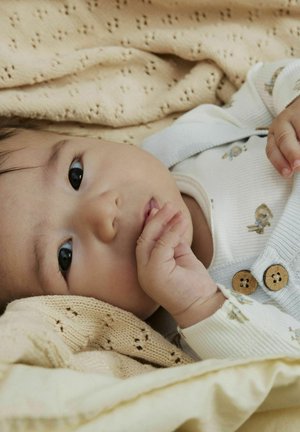 Bebé tumbado sobre una manta beige texturizada, vistiendo un body blanco con textura de canalé con adorables estampados de animales y tirantes azul claro con botones de madera.